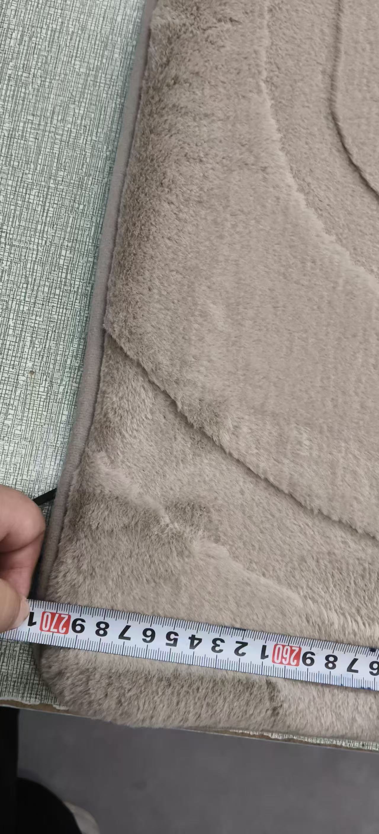 A close-up of a beige plush or fleece fabric product being measured with a tape measure, with a hand holding the tape at the edge.