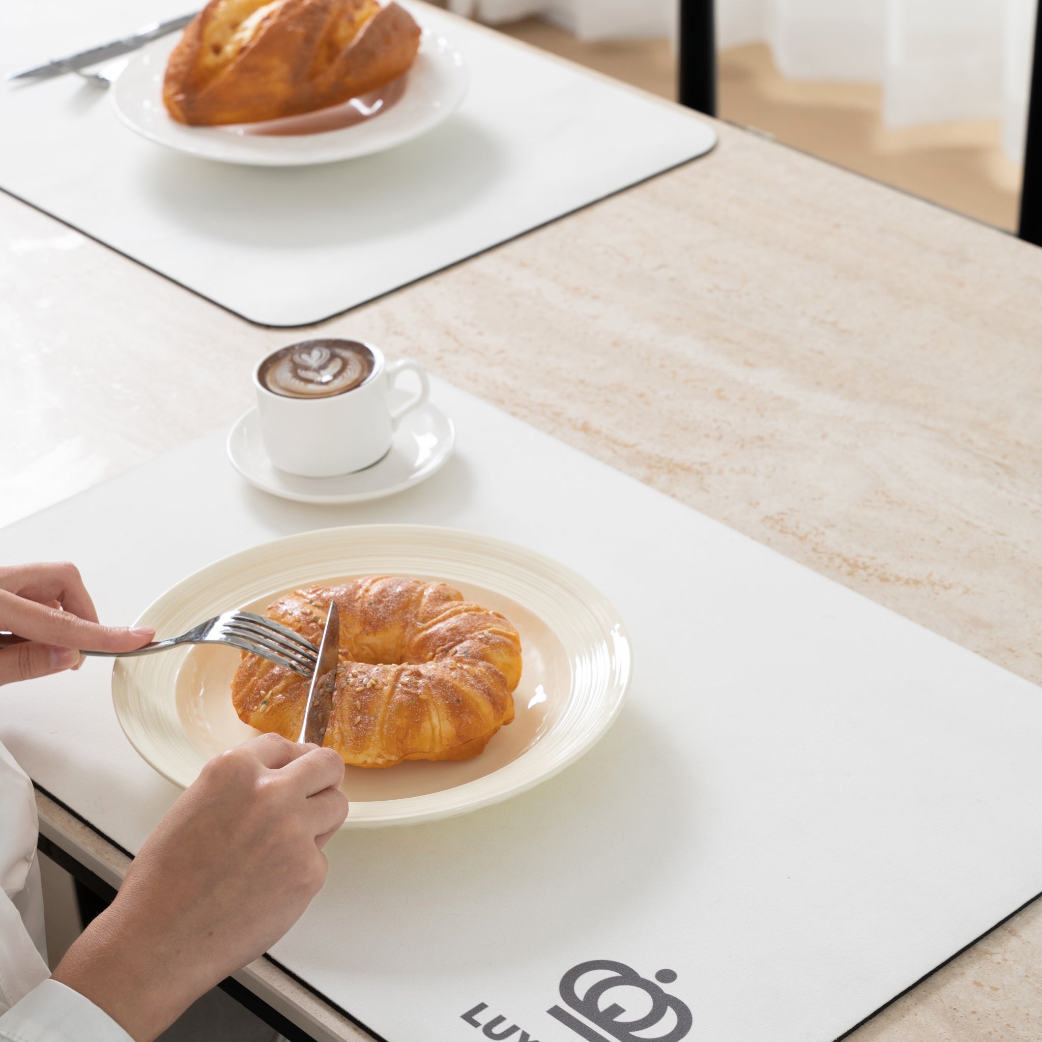 Person cutting a croissant with a knife and fork on a white plate, with a cup of coffee in the background.