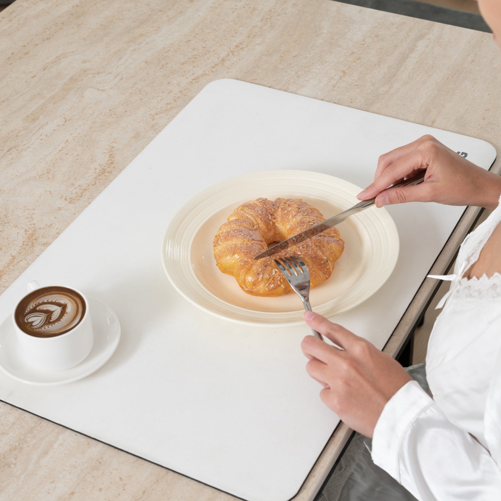 Person cutting a pastry on a white plate with a knife and fork, next to a cup of coffee on a white mat.