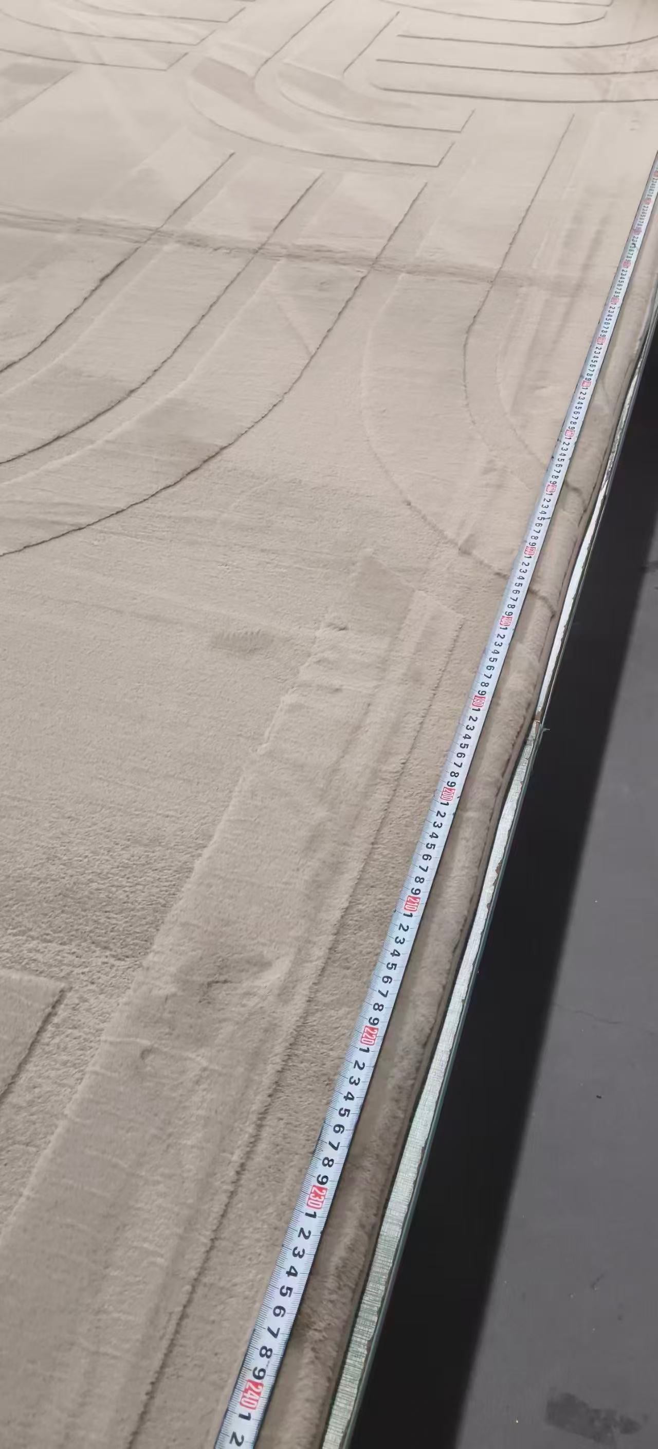 A close-up view of a light beige textured carpet or rug with a white measuring tape laid along one edge to show the length.