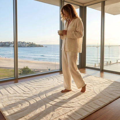 Woman in beige outfit standing in a room with large windows overlooking a beach.