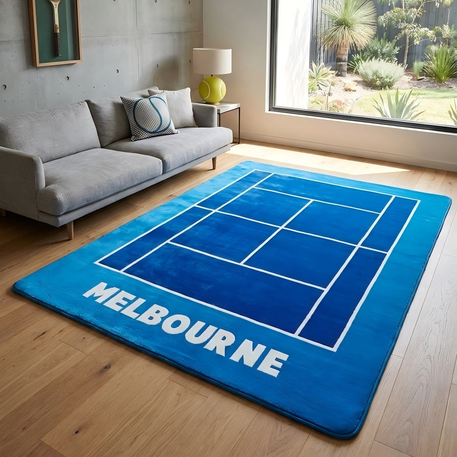 A blue rectangular area rug with a tennis court line design and the word 'MELBOURNE' in large white letters, shown on a wood floor in a modern living room next to a gray sofa and large window.