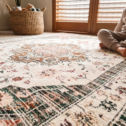 A low-angle lifestyle shot of a vintage-style area rug with a distressed medallion pattern in cream, rust, green, and pink. A child is sitting on the right edge of the rug, and a woven basket is visible in the background near wooden doors.