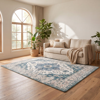 A large rectangular area rug with a traditional medallion pattern in muted blue, beige, and cream tones, styled on a light wood floor in a bright, modern living room with a beige sofa and potted plants.