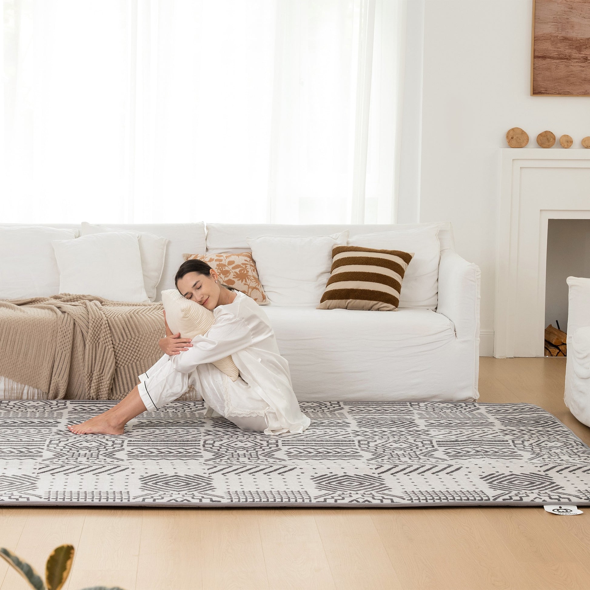Woman sitting on a patterned rug in a living room with a white couch and decorative pillows.