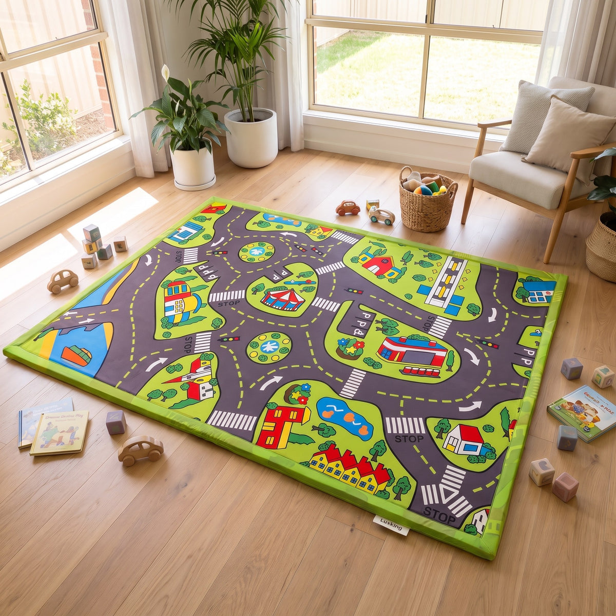 A large children's road-play rug with a green border and illustrated winding roads, cars, houses, trees, and traffic lights, shown on a wooden floor in a bright playroom with toys, plants, and a chair nearby.