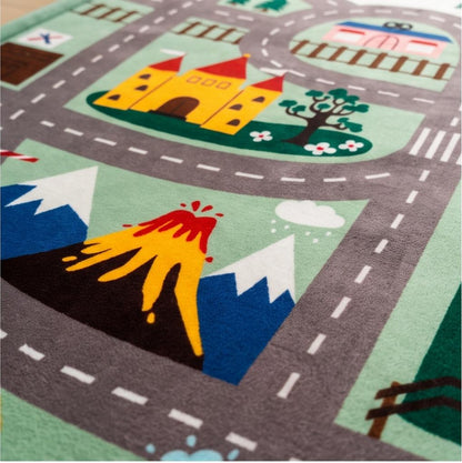 A close-up of a children's play rug featuring a colorful road-travel scene with gray winding roads, a yellow castle, blue and white mountains, an erupting volcano, trees, and small train tracks.