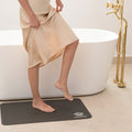 Person stepping onto a black mat in a bathroom with a white bathtub and gold fixtures.