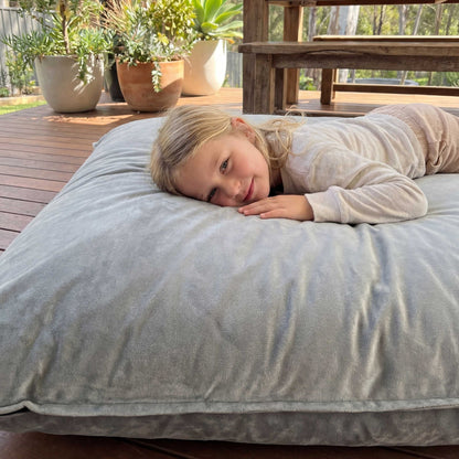Person lying on a large cushion outdoors with plants and a wooden table in the background