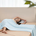 Woman lying on a bed with a light blue blanket, surrounded by a neutral-colored room.