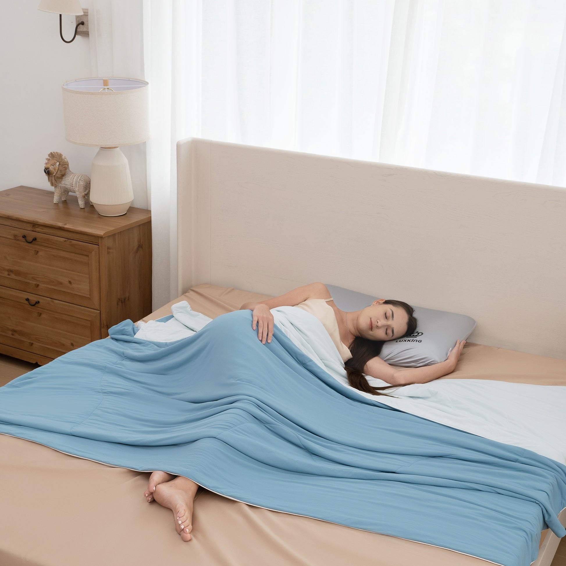 Woman sleeping under a blue blanket in a bedroom with a wooden nightstand and lamp.